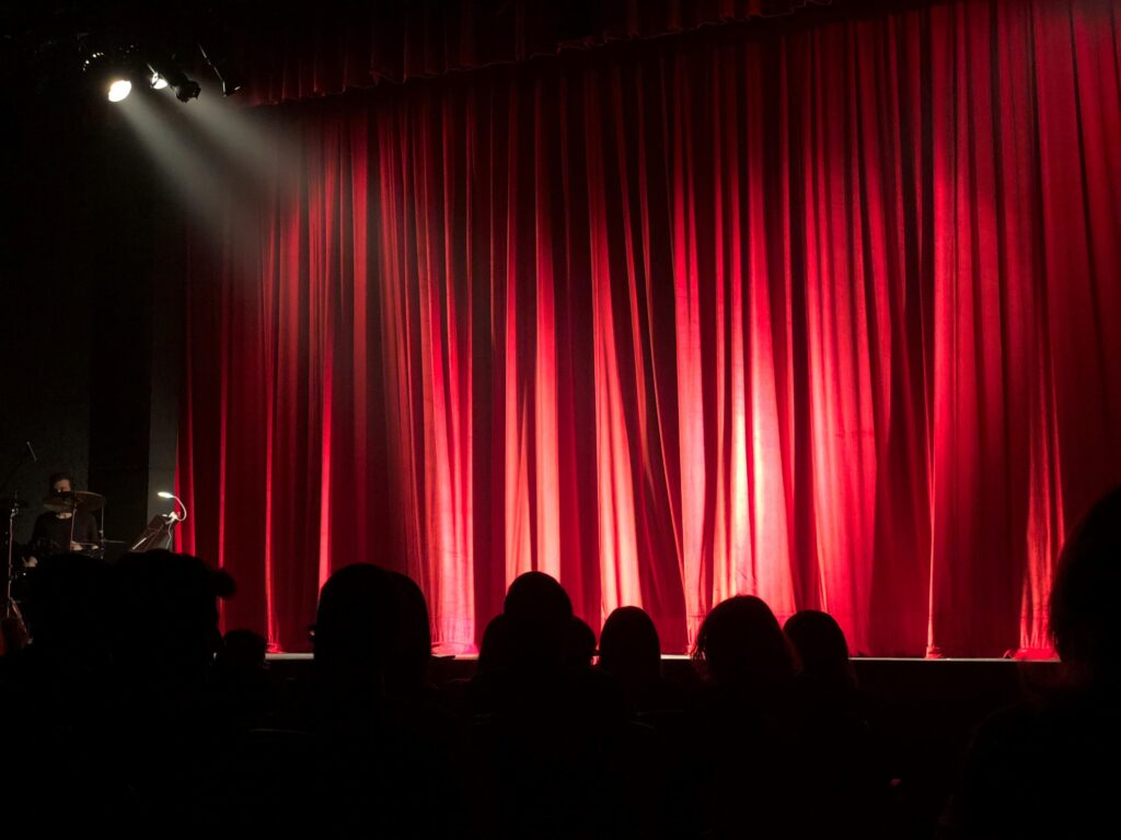 Dimly lit theater stage with red curtains and audience silhouettes under spotlights.